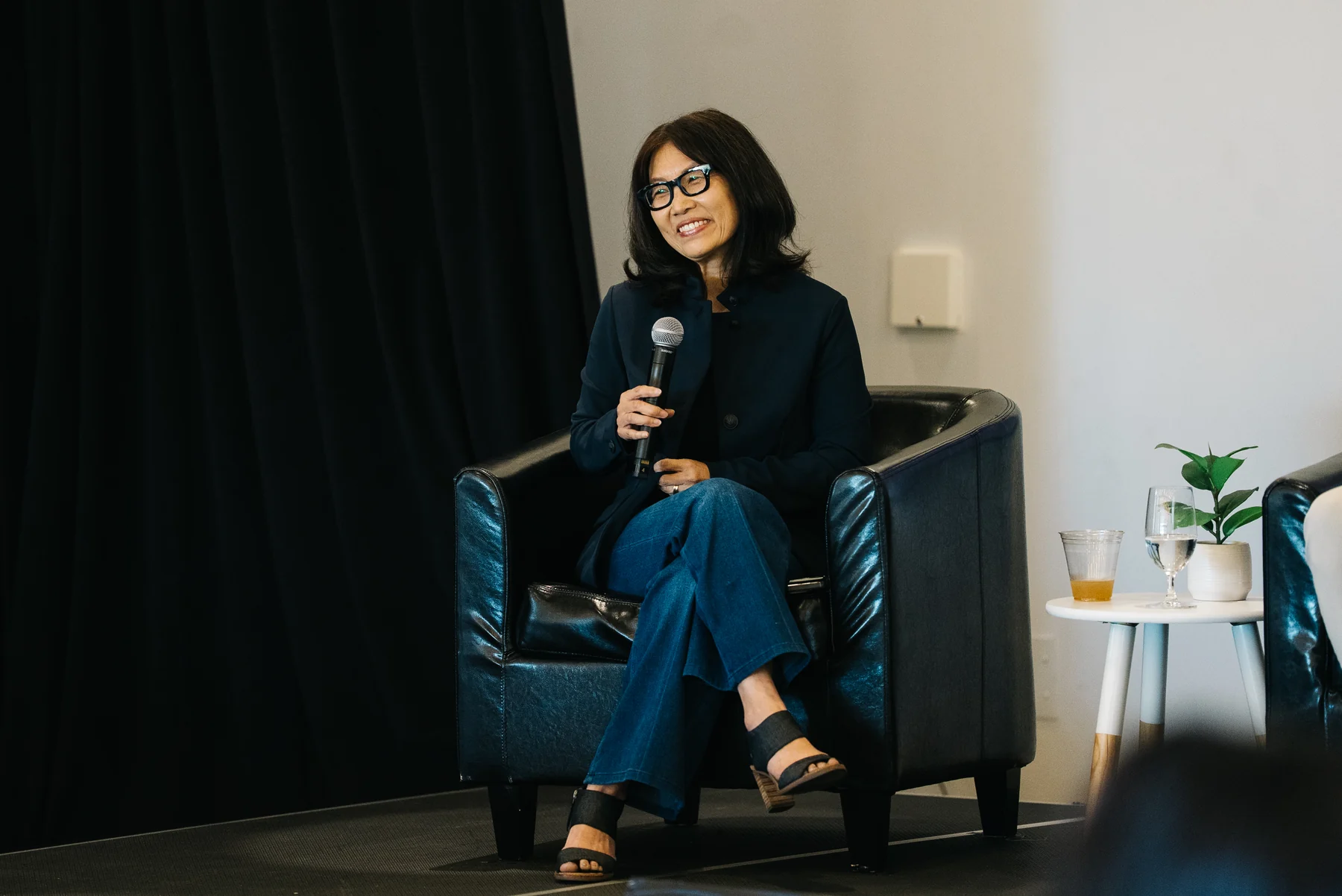 A panelist with glasses smiles while holding a microphone, seated in a black armchair.