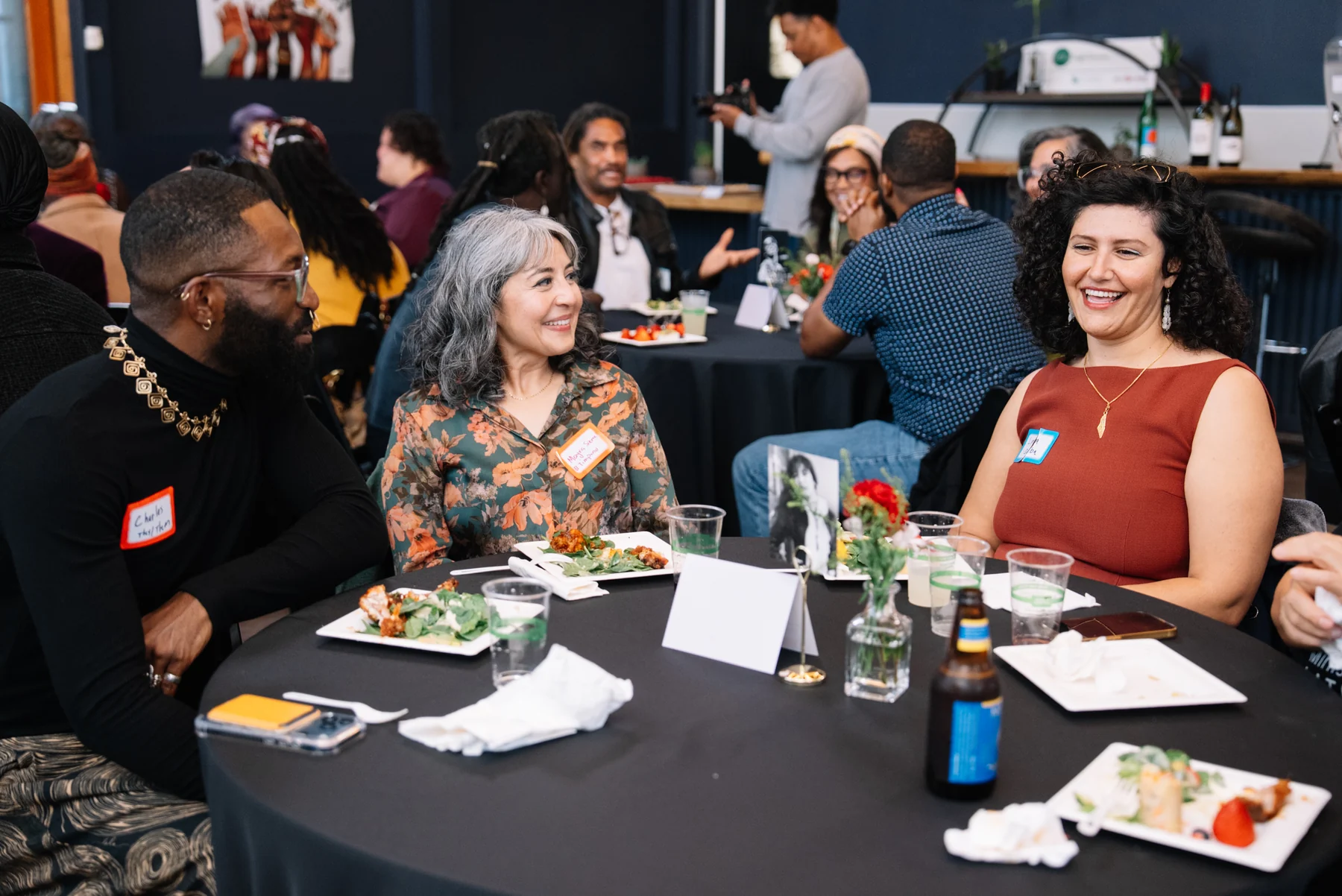 Three guests smile and talk around a round table with food and flowers.