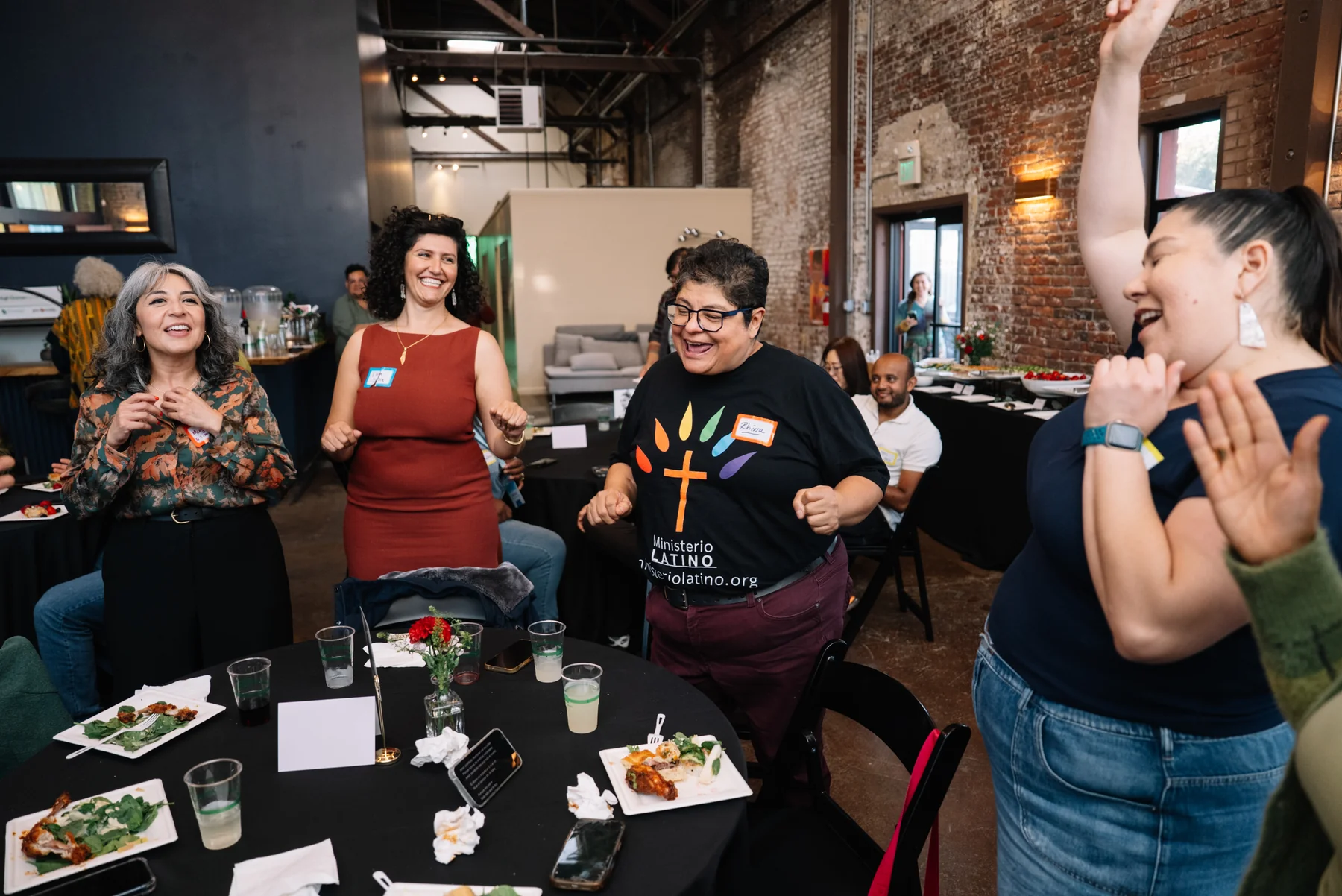 Guests laugh and raise their hands around a table in a sunlit brick-walled room.