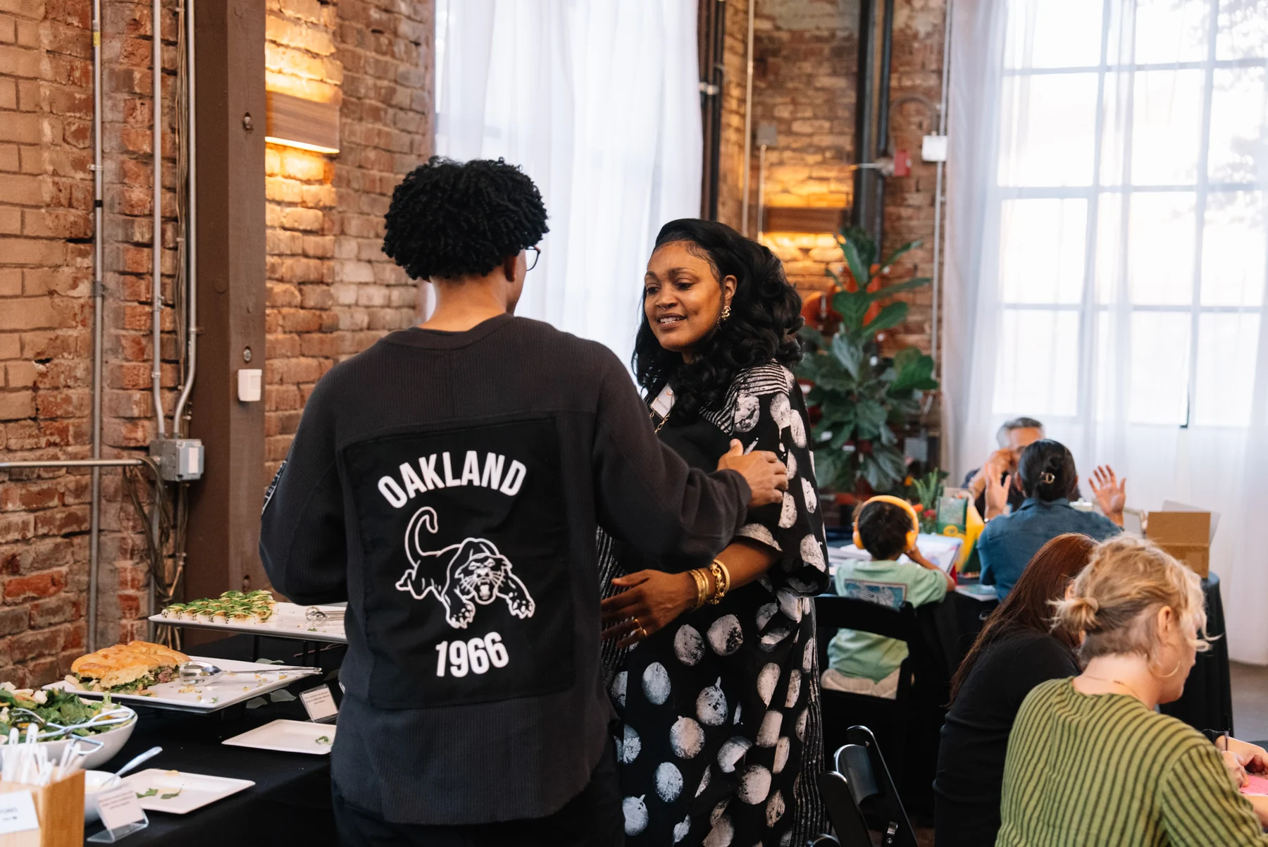 Two attendees speak together beside a buffet table in a sunlit brick-walled room.