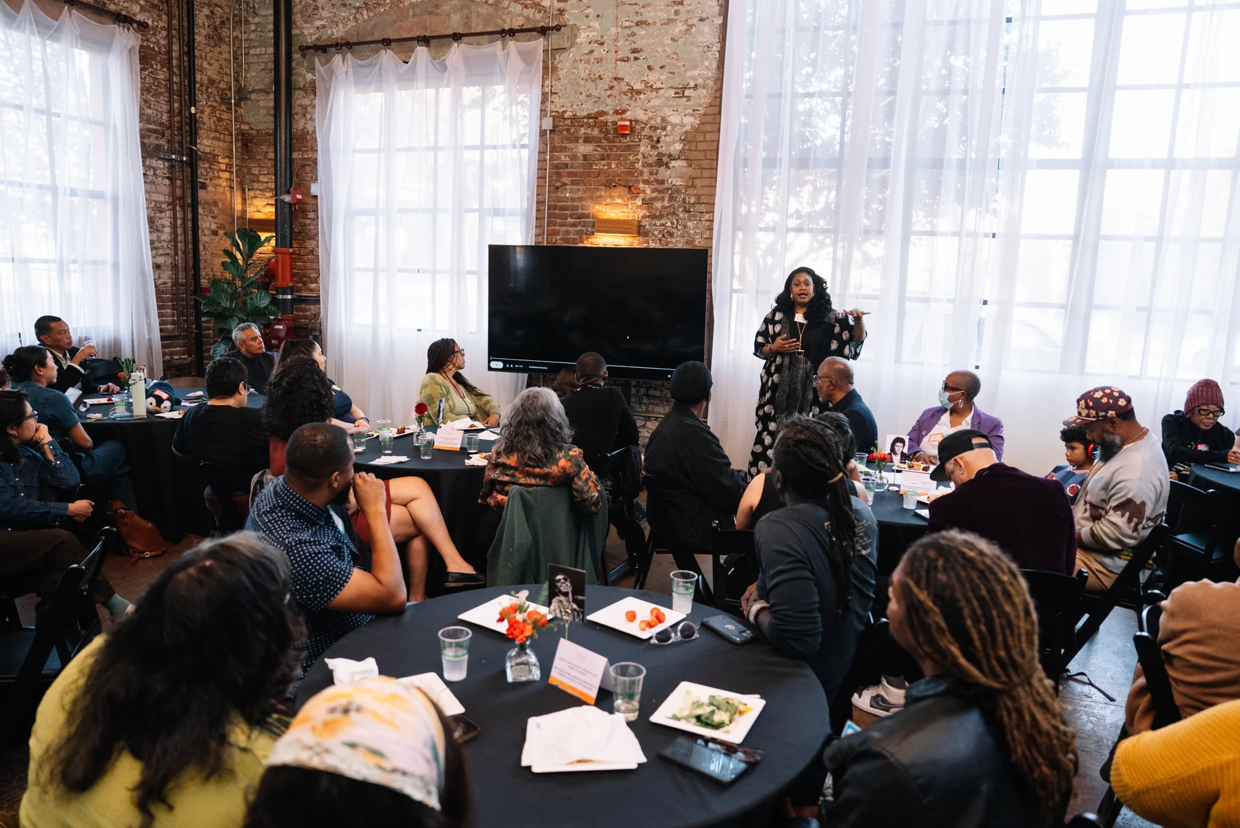 Guests seated at round tables listen to a standing speaker beneath tall windows.