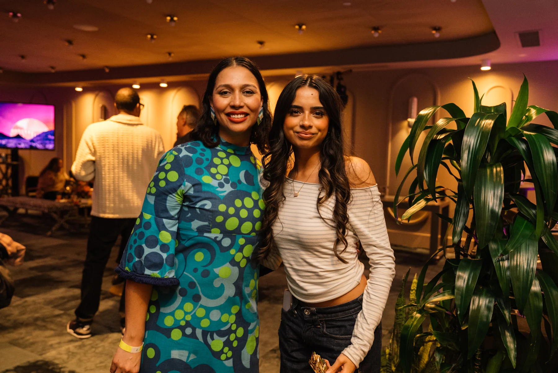 Two guests stand together beside a leafy plant at the celebration.