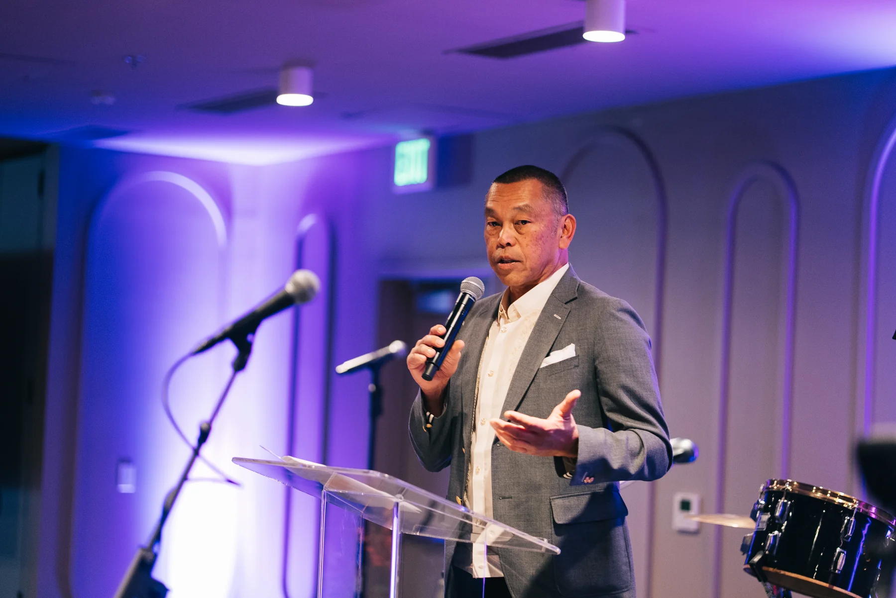 A speaker holding a microphone gestures from a clear lectern in purple light.
