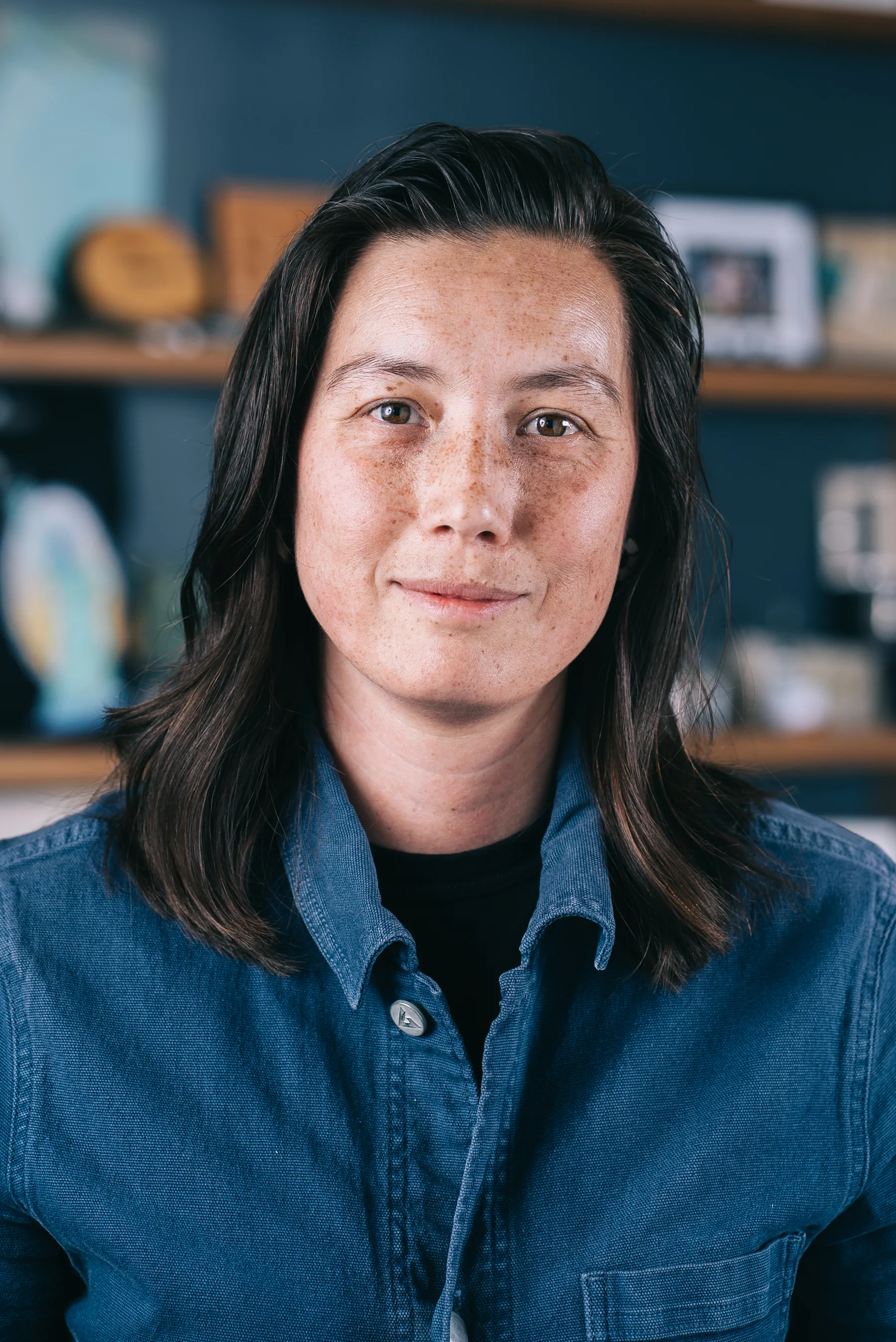 A participant with dark shoulder-length hair wears a blue shirt in front of teal shelves.