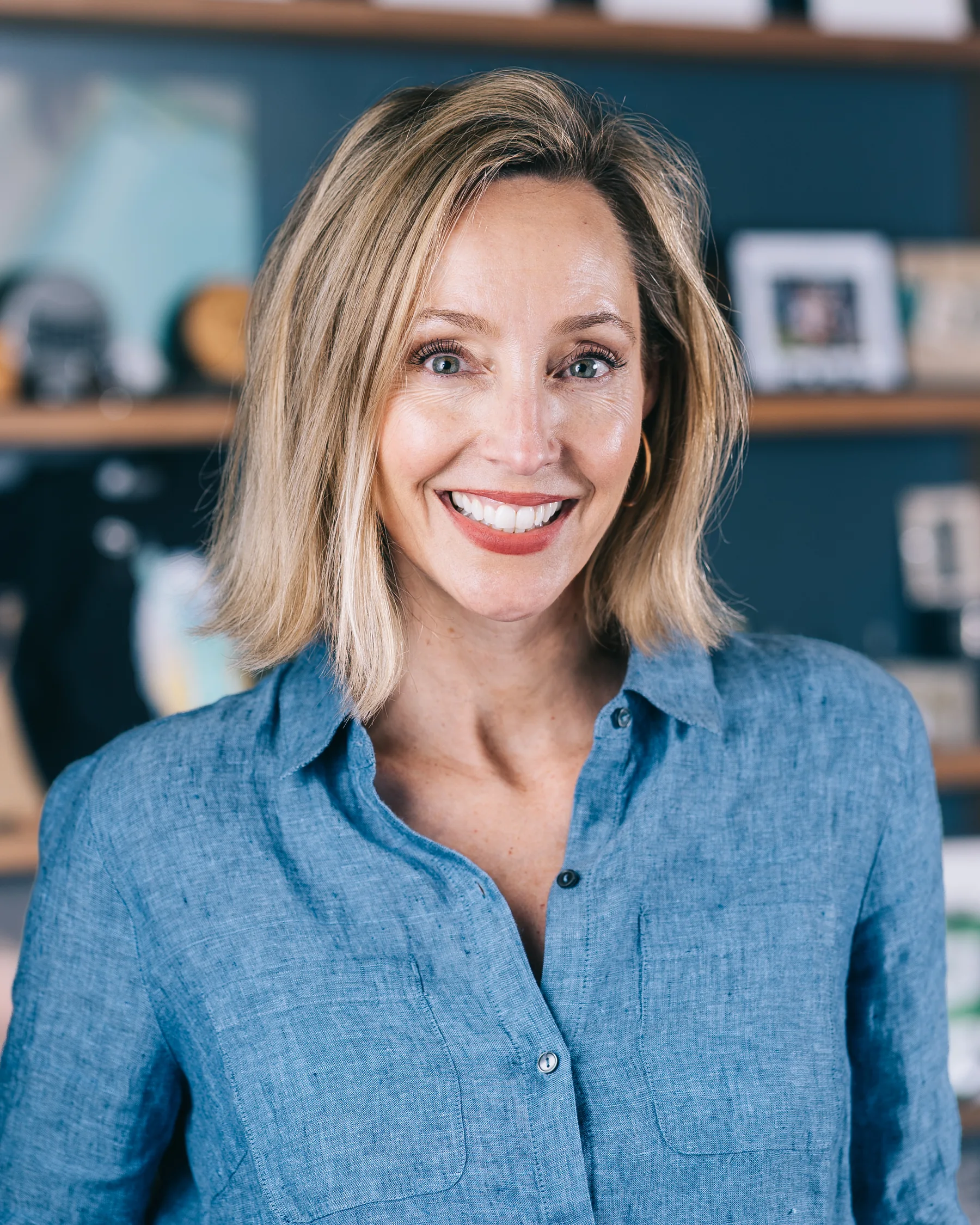 A smiling participant with shoulder-length blonde hair wears a blue shirt in front of teal shelves.
