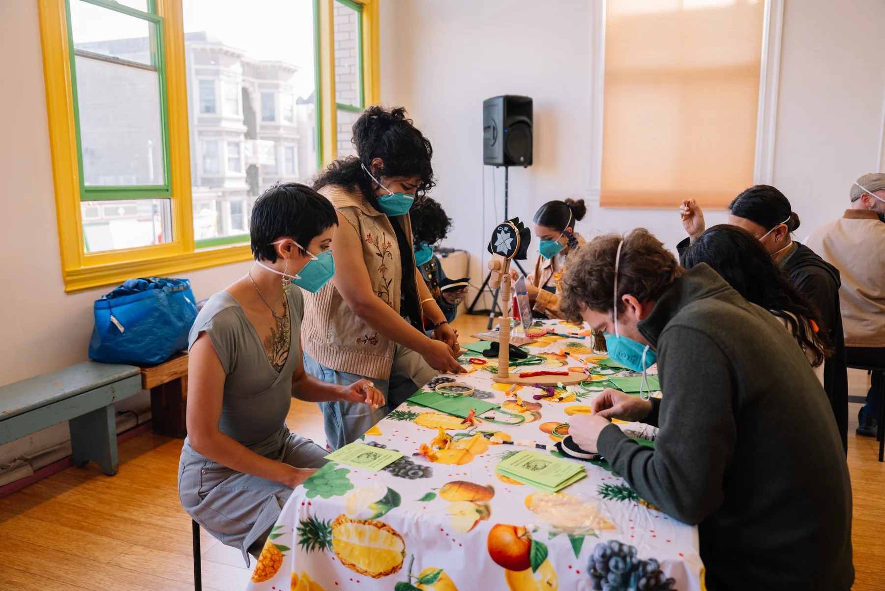 People work with embroidery hoops and thread around a long table beside yellow-and-green windows.