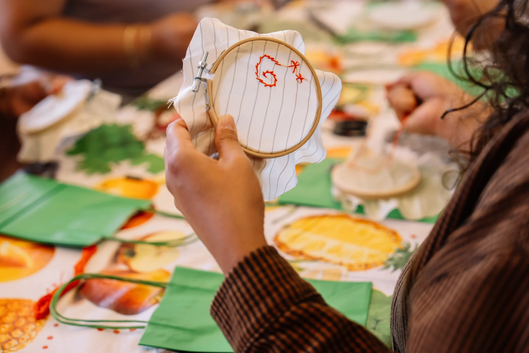 Hands hold an embroidery hoop with red stitching above a table of fabric and craft supplies.