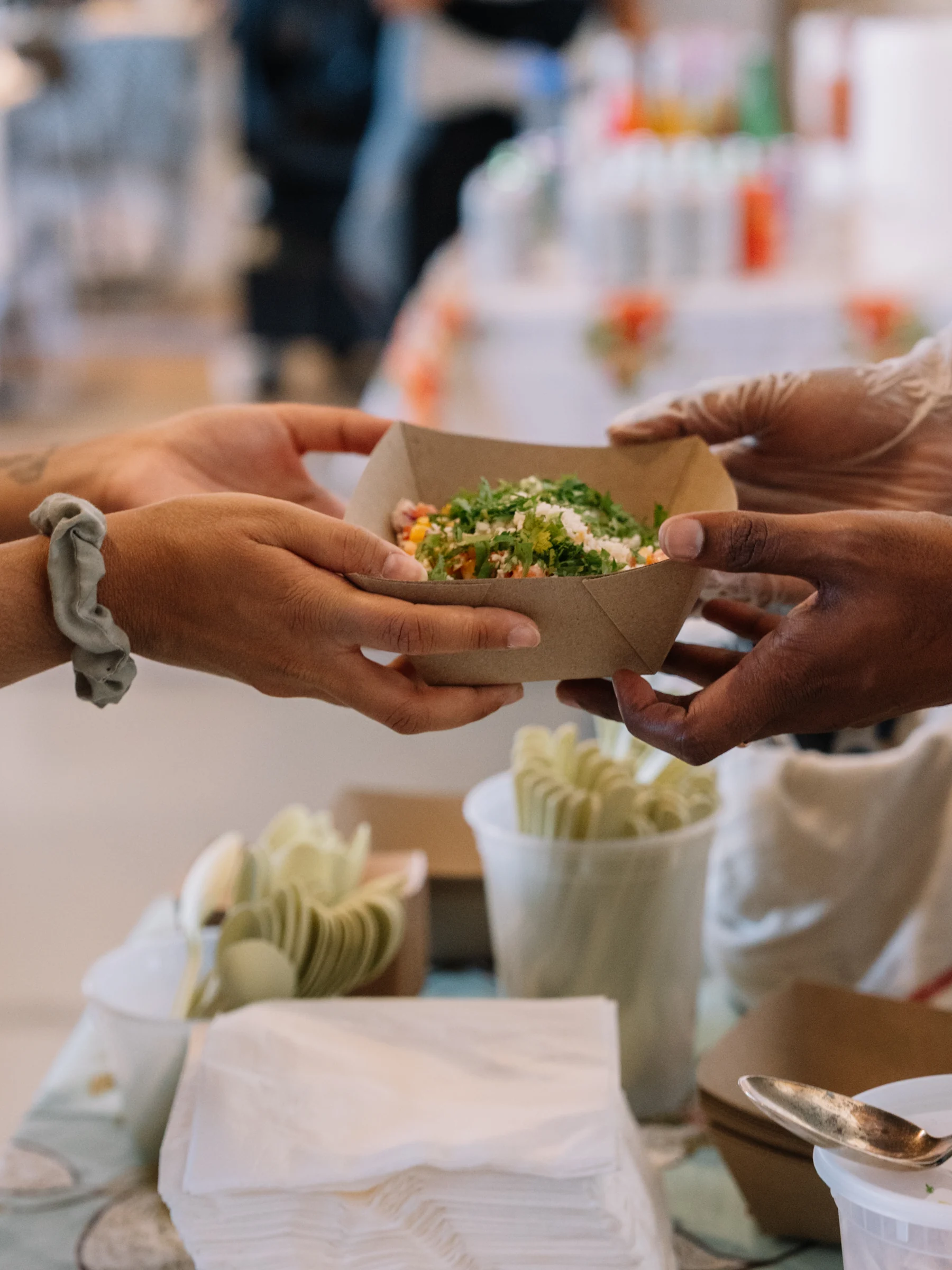 Two pairs of hands pass a paper tray of food across a serving table.