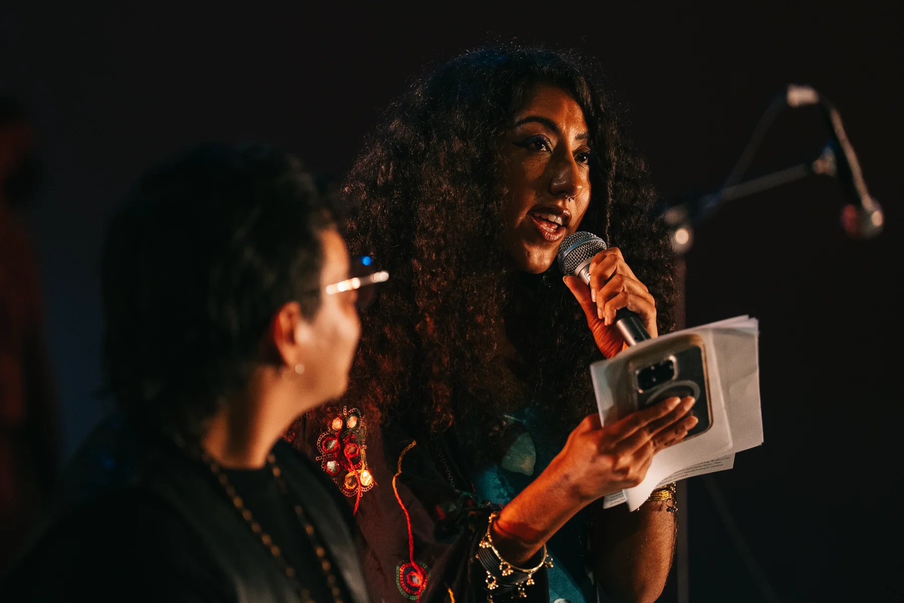 A speaker holds a microphone, papers and a phone on stage, with another person in the foreground.