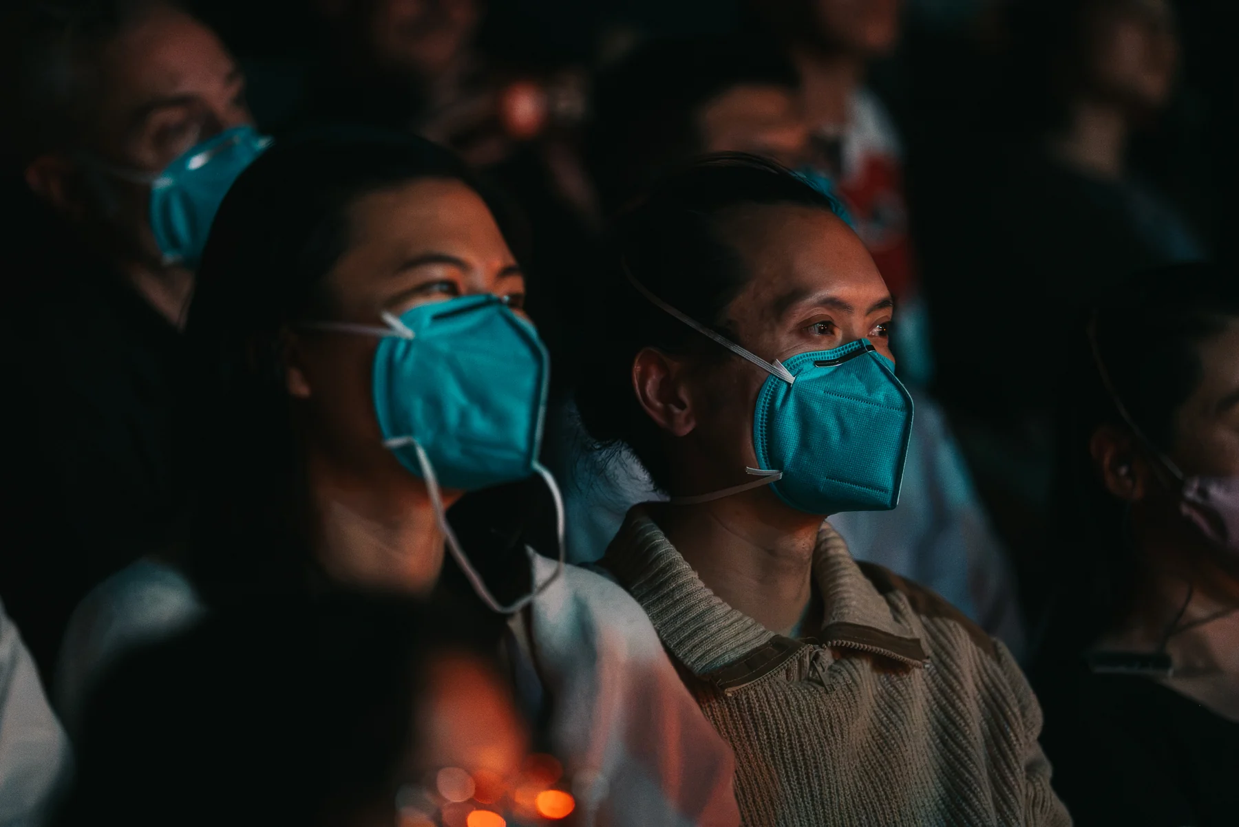 Two audience members in teal face masks sit among others in a darkened theater.