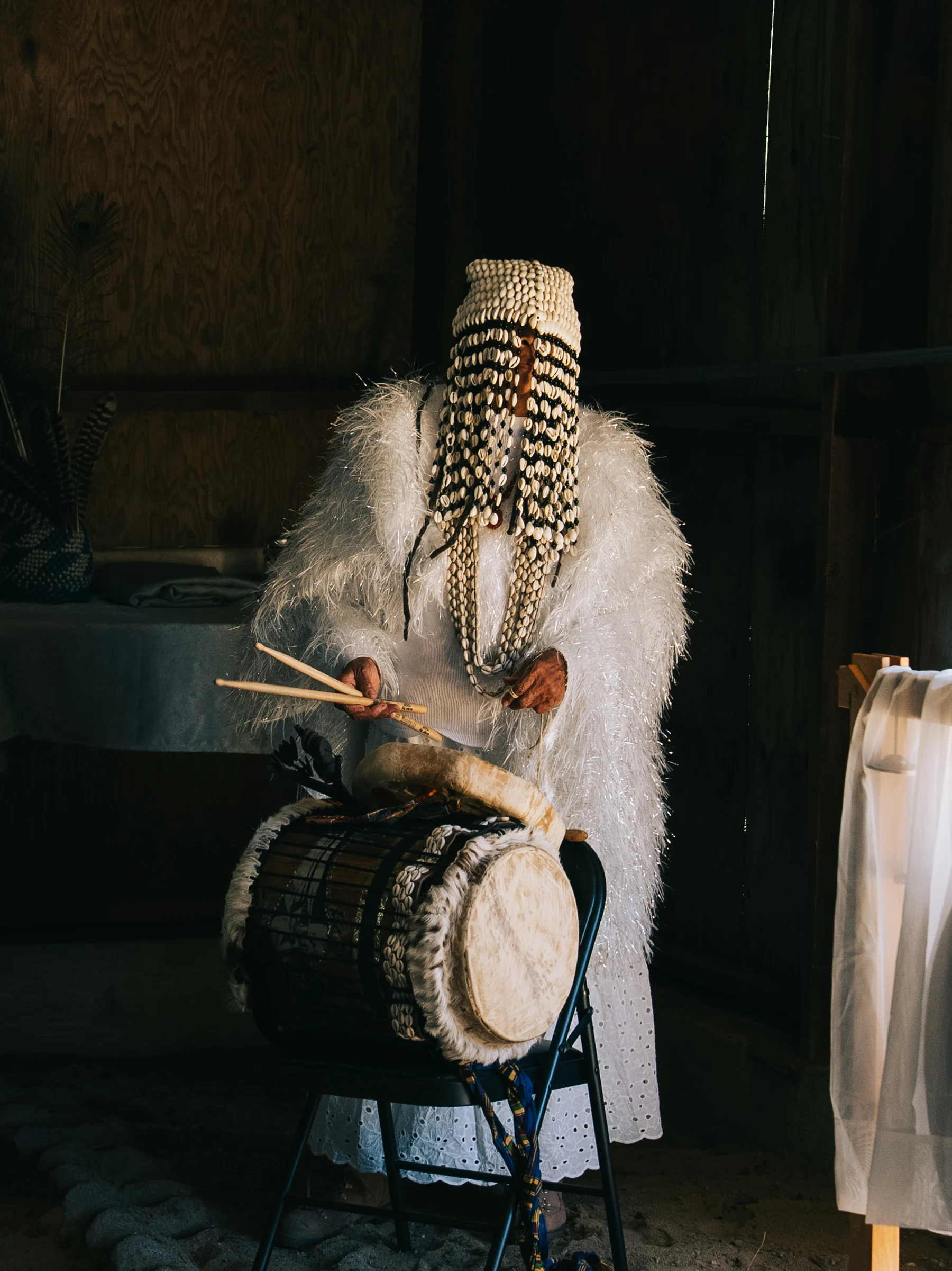 A seated drummer with long braids plays a hand drum in a shaft of light.
