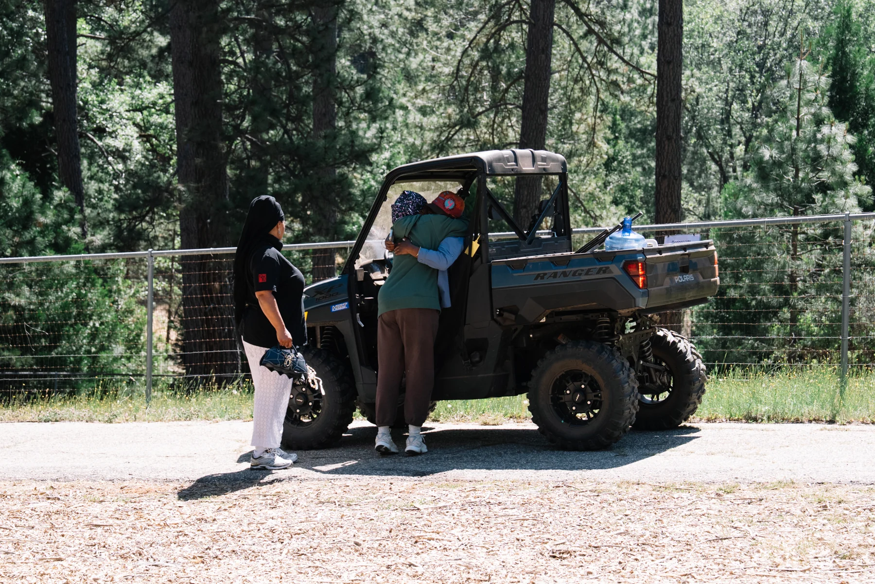 Two people embrace beside a utility vehicle among tall trees, while another person stands nearby.