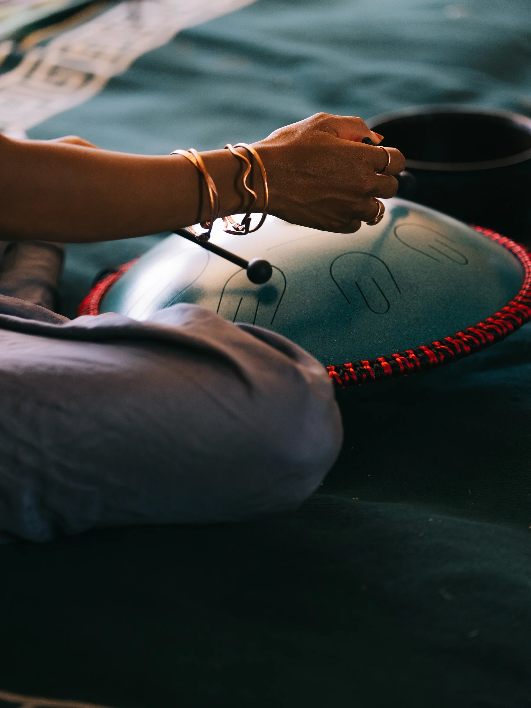 Hands hold mallets above a teal handpan resting in a seated participant’s lap.
