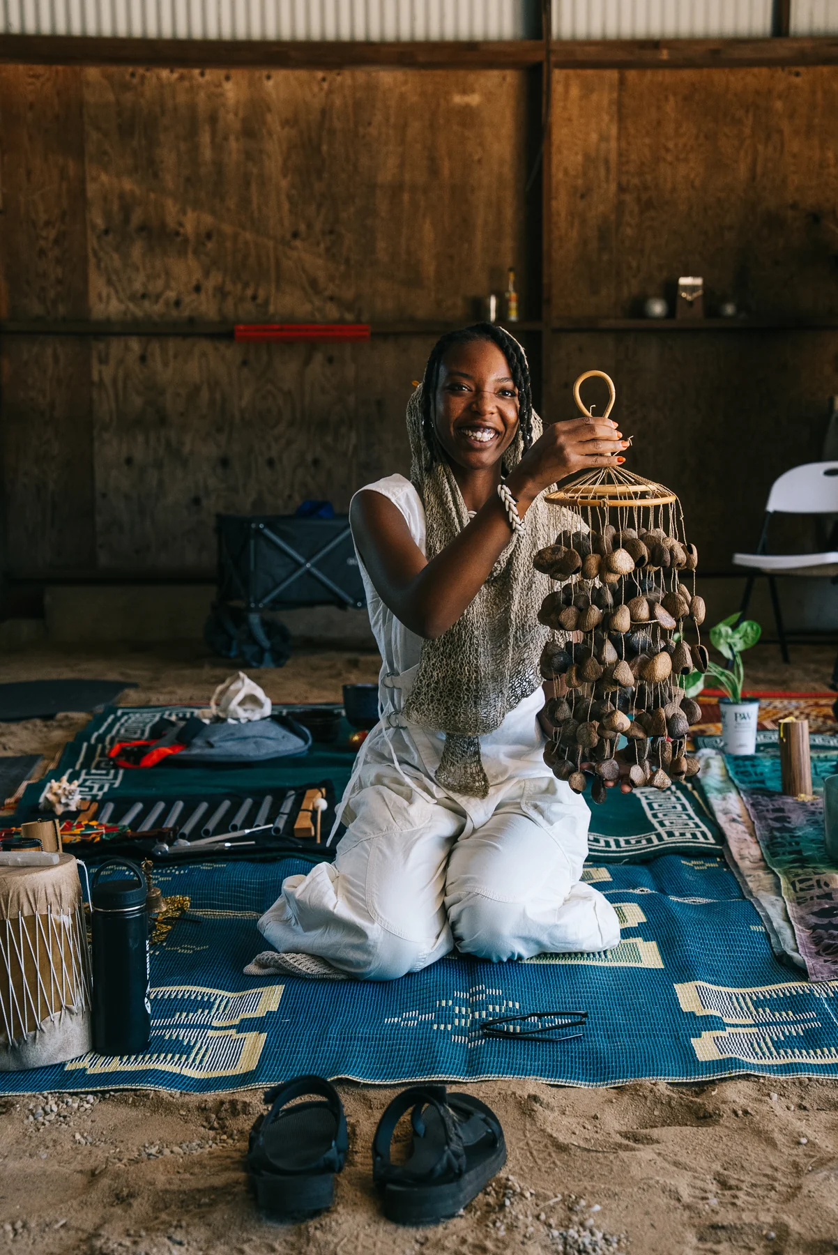 A smiling participant holds a hanging percussion instrument while seated on a rug.