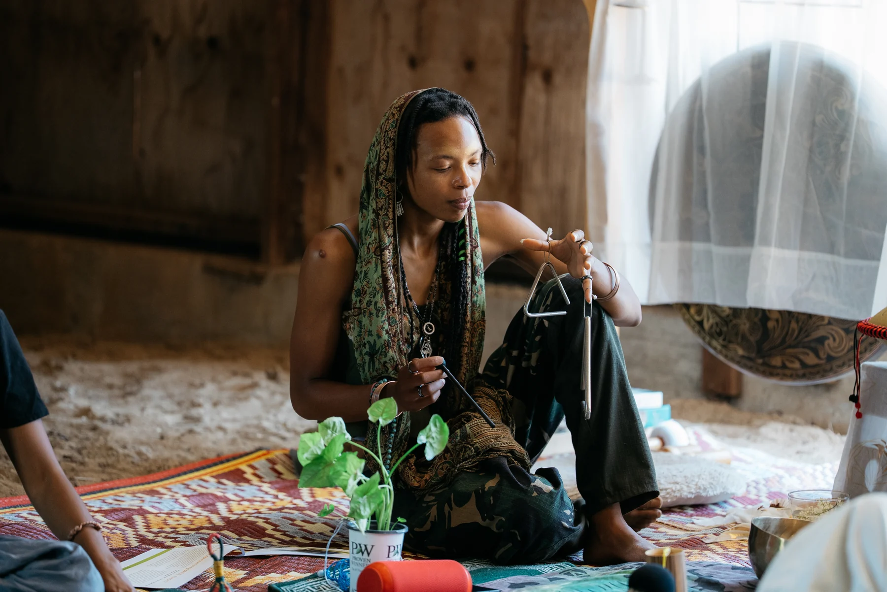A participant seated on patterned rugs holds a triangle beside other instruments.