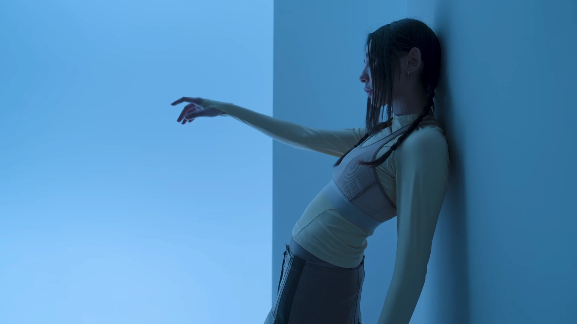A woman leans against a blue-lit wall with one arm extended.