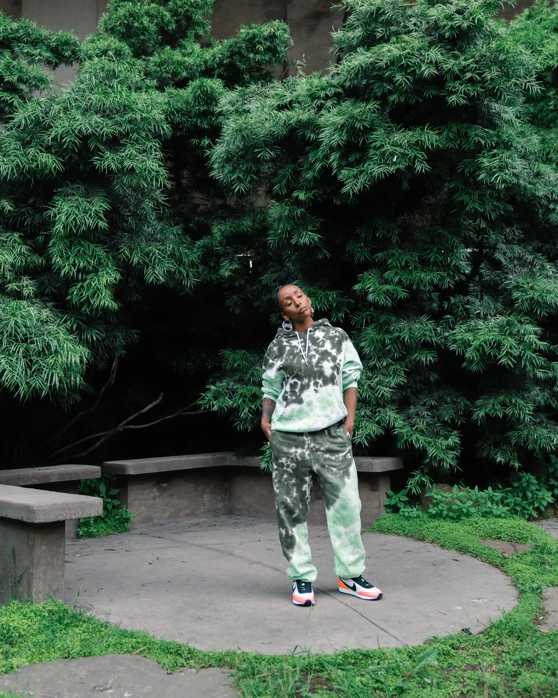 ASTU stands in a green patterned outfit on a garden path beneath dense trees.