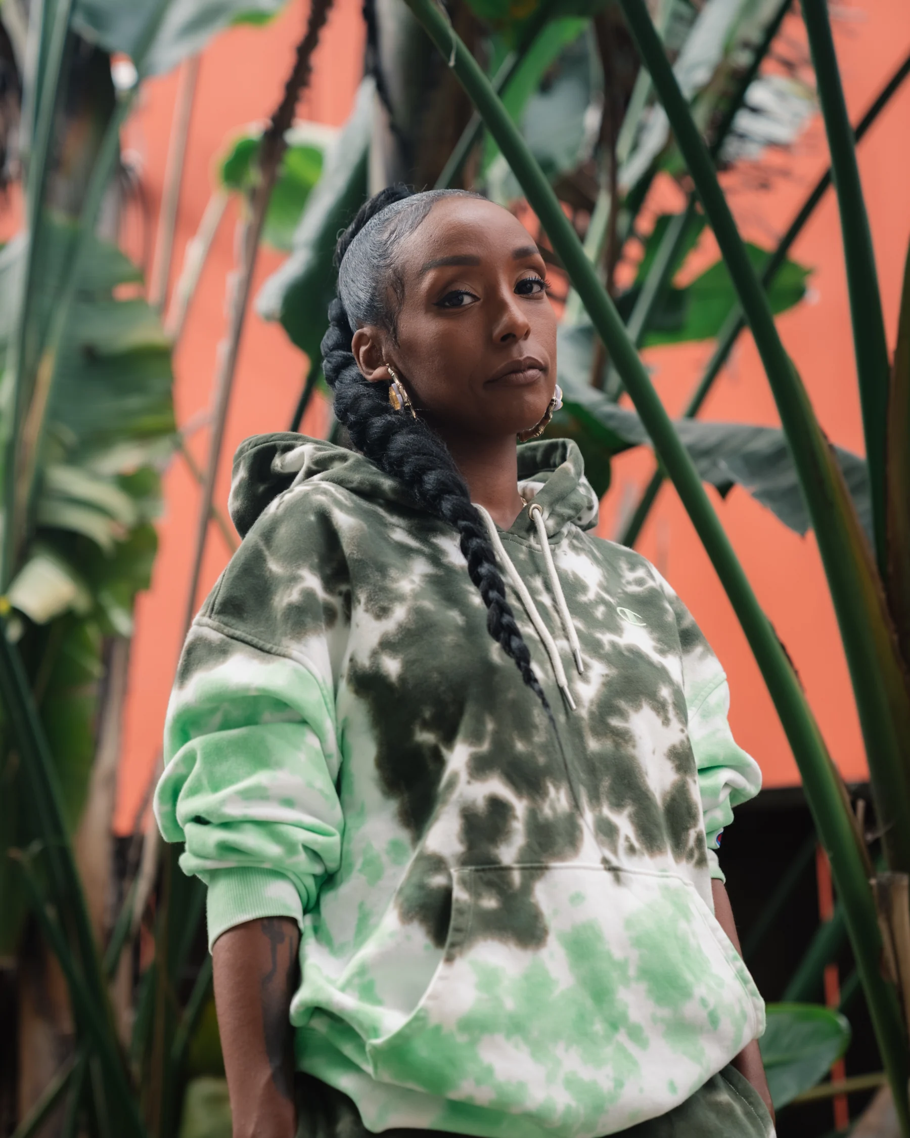 Portrait in a green-and-white hoodie, framed by tall leaves against a coral wall.