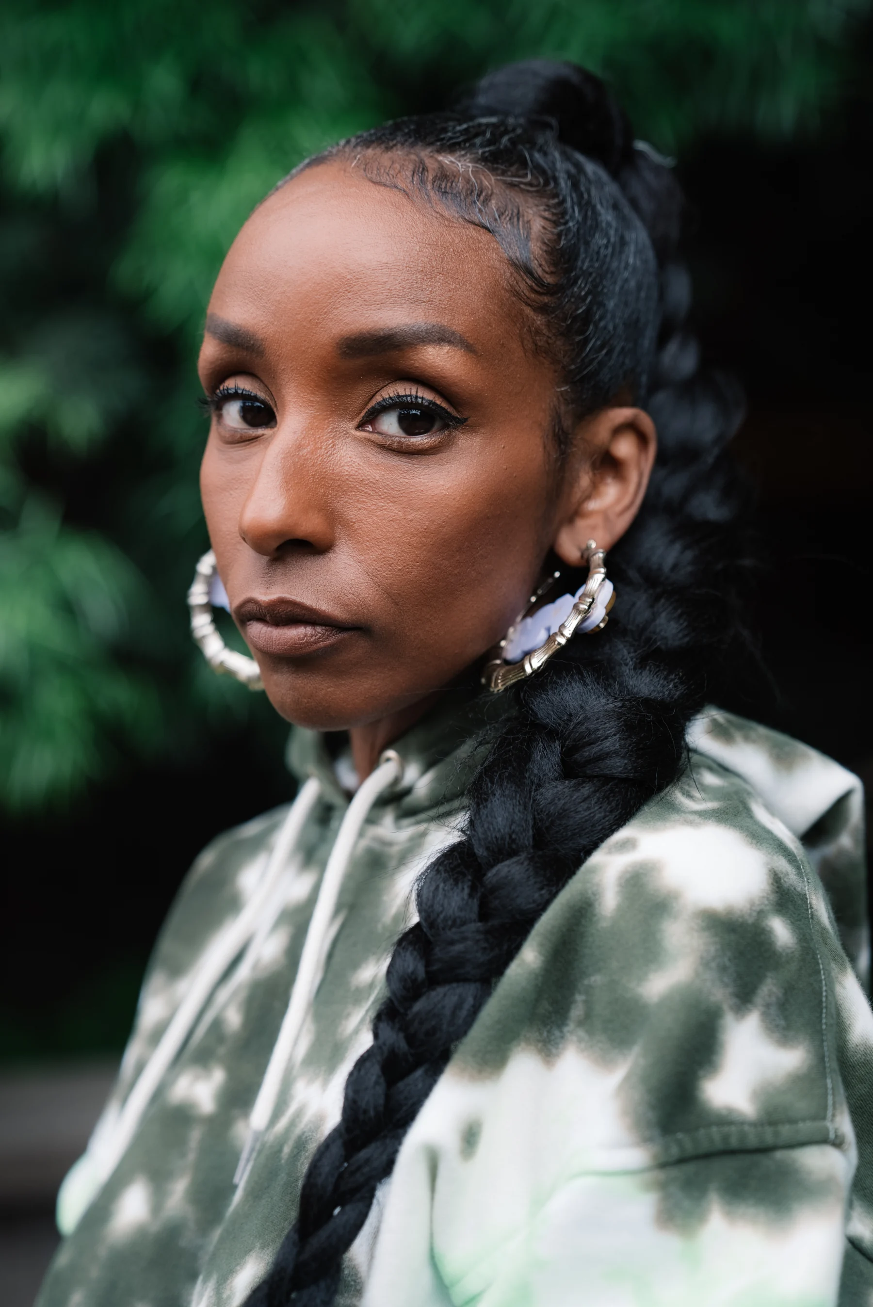 Close portrait with a long braid and large earrings against green foliage.