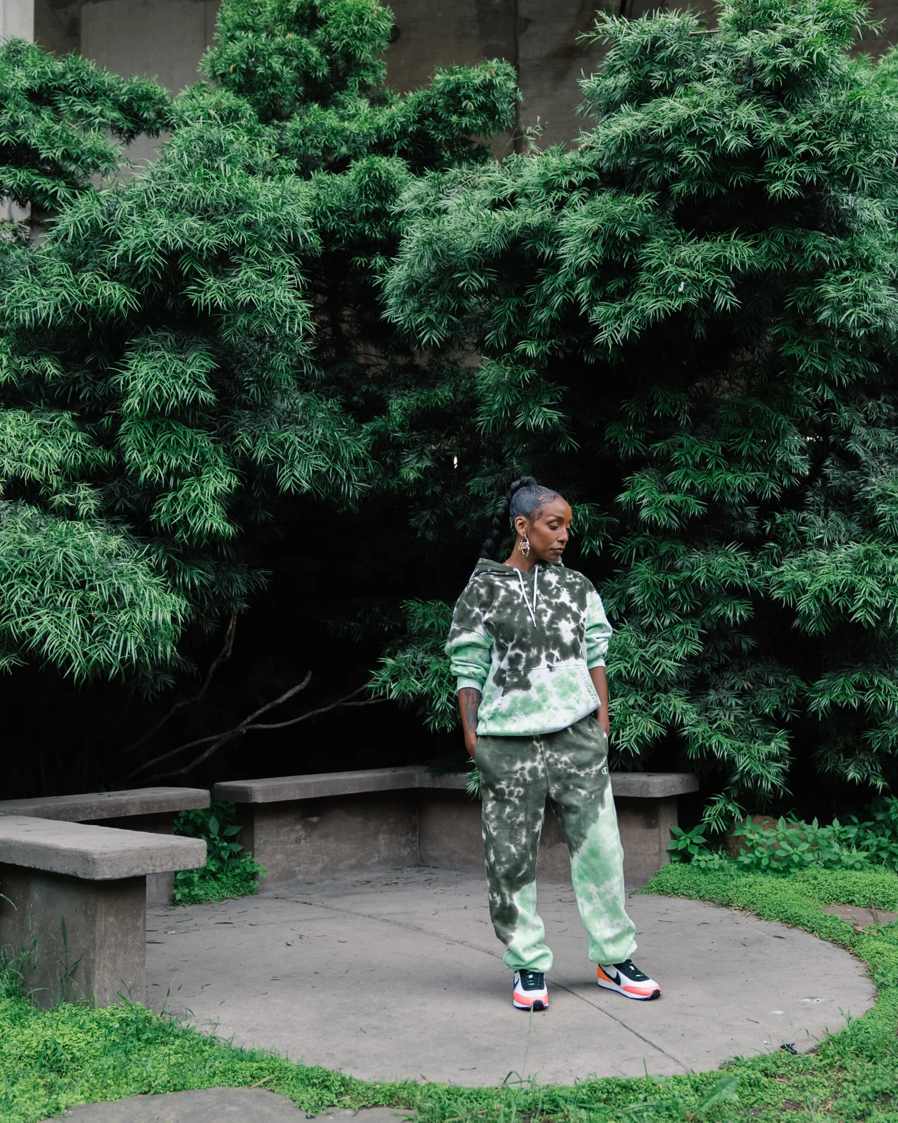 Full-length portrait in a matching green-and-white outfit beside concrete benches and dense greenery.