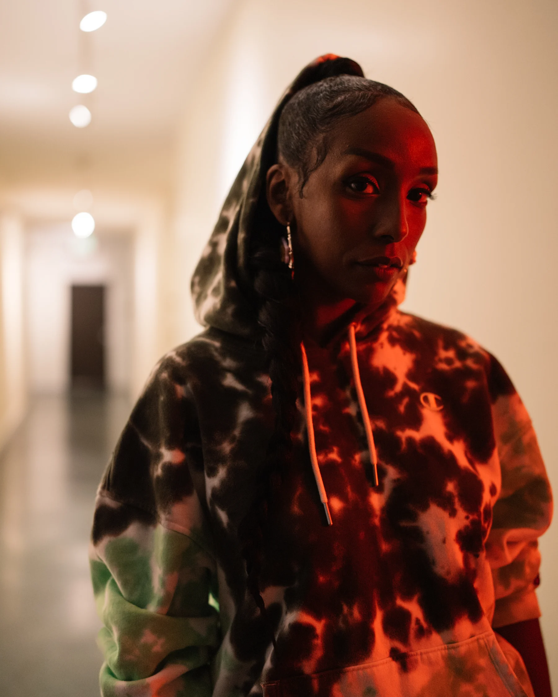 Portrait in a light hallway with red light falling across a patterned hoodie.