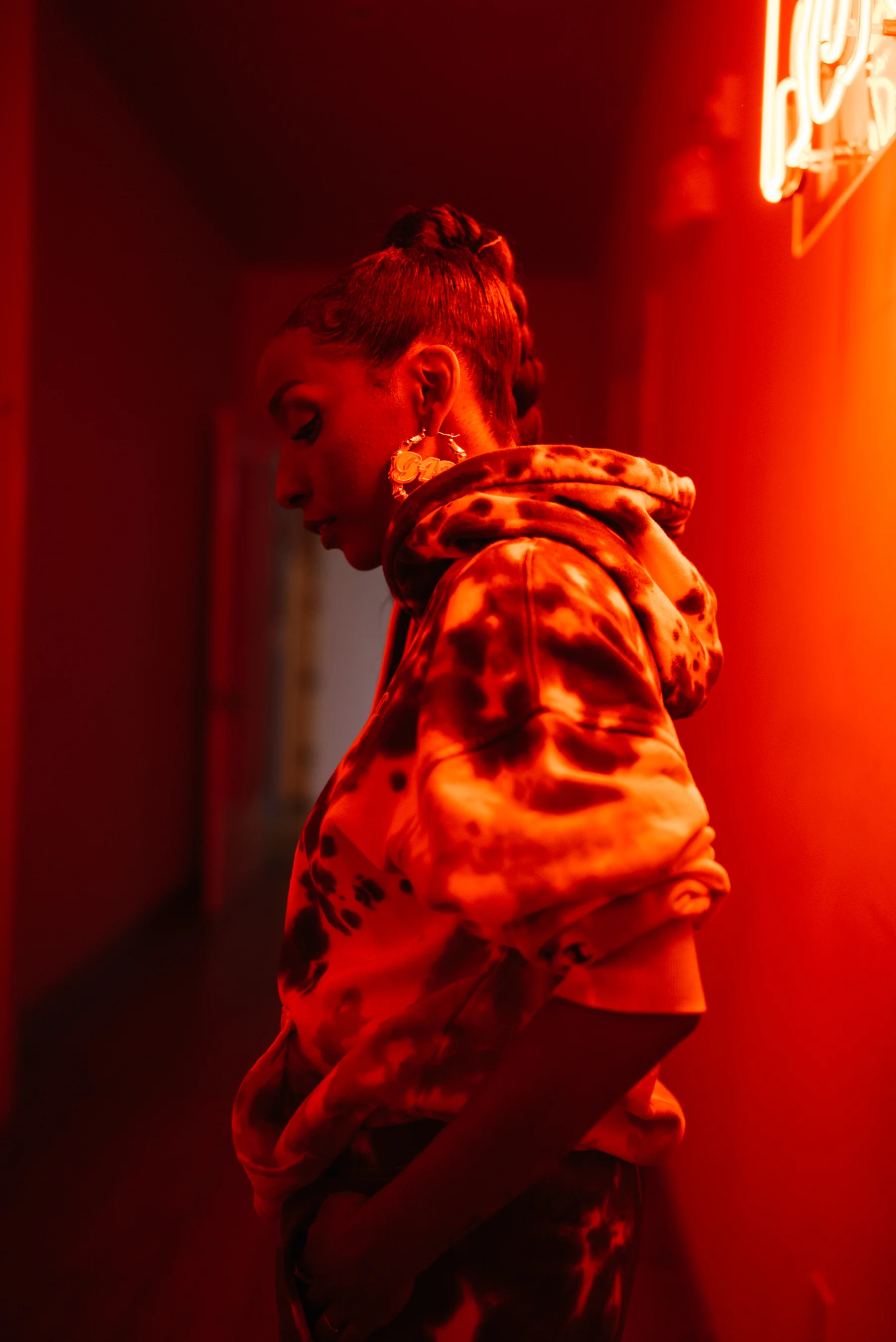 Side-profile portrait in a patterned hoodie beneath warm red neon light.