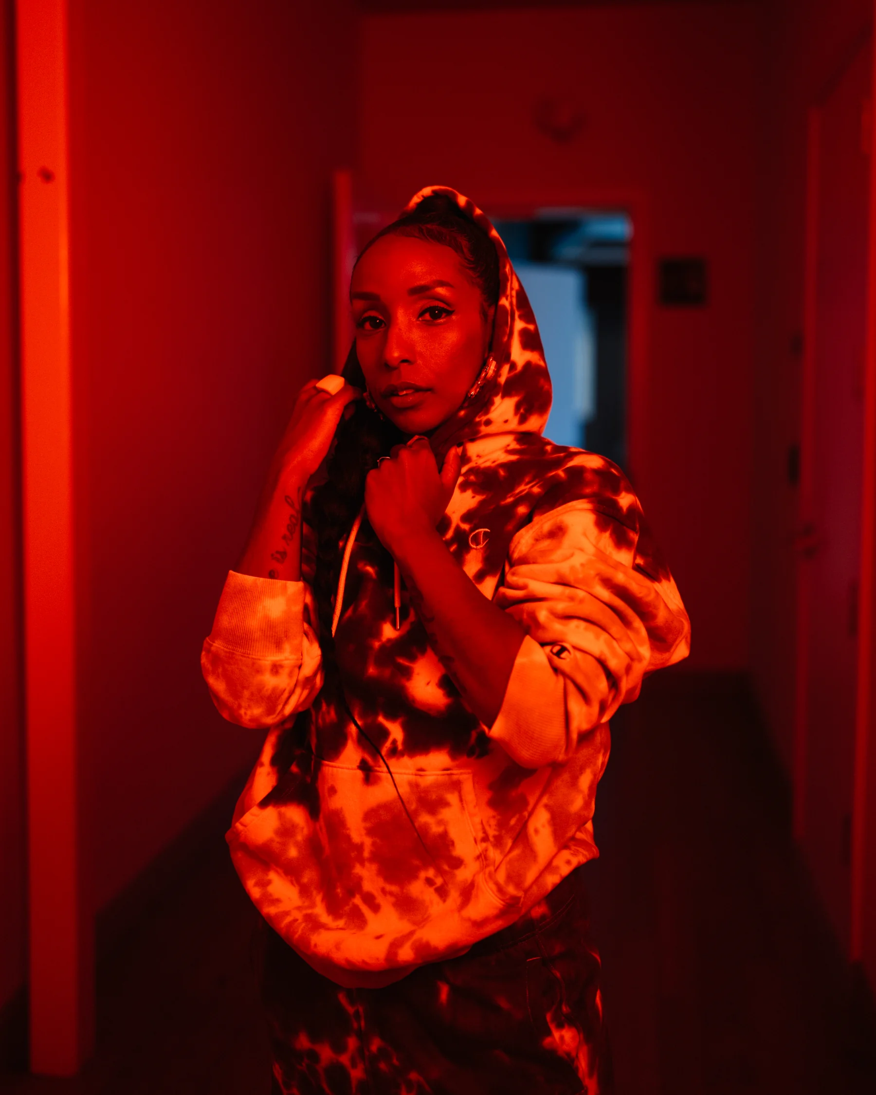 Portrait with both hands holding a raised hood in a red-lit corridor.