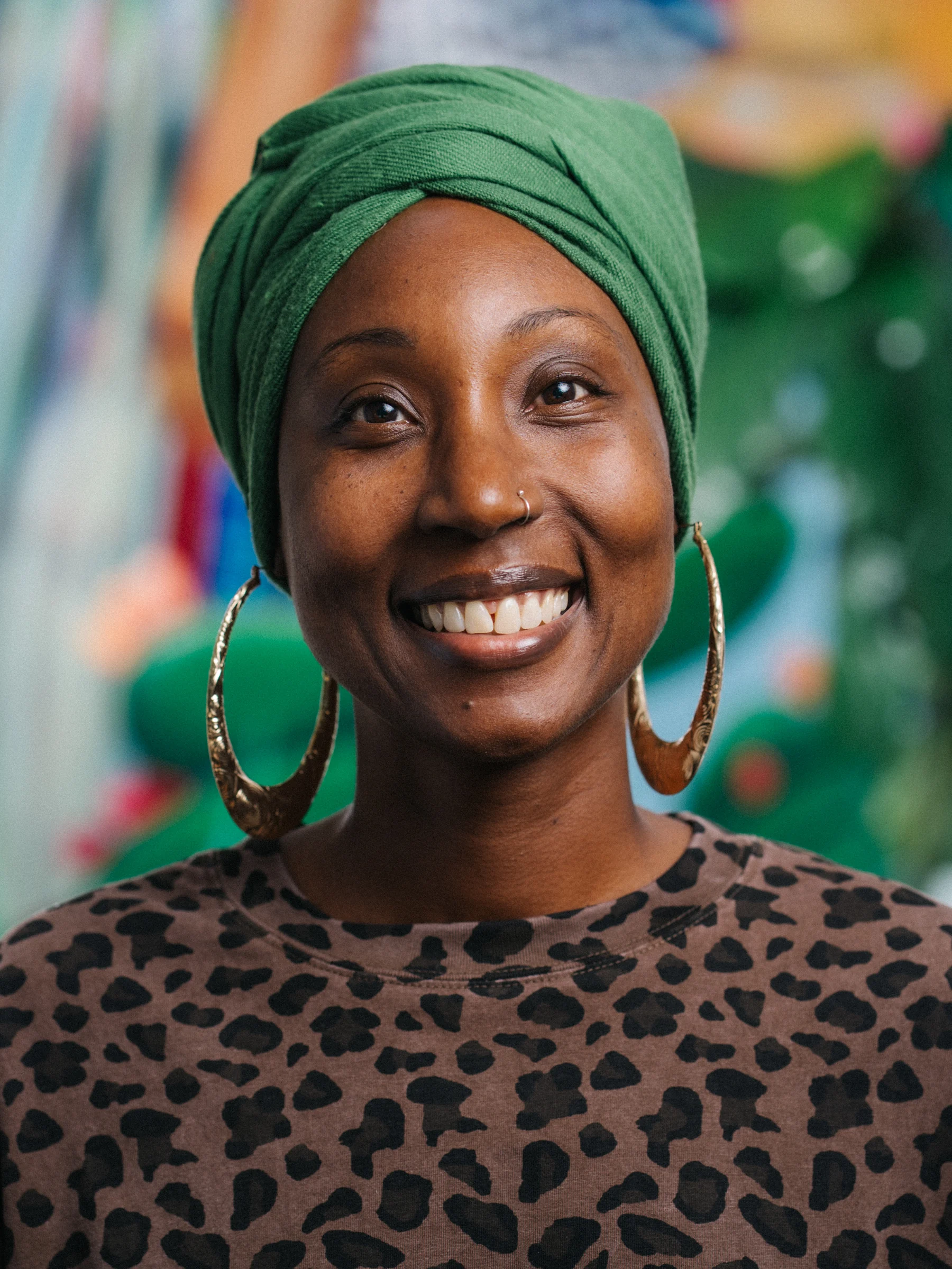 A smiling subject in a green head wrap and patterned top against a softly blurred, colorful mural.
