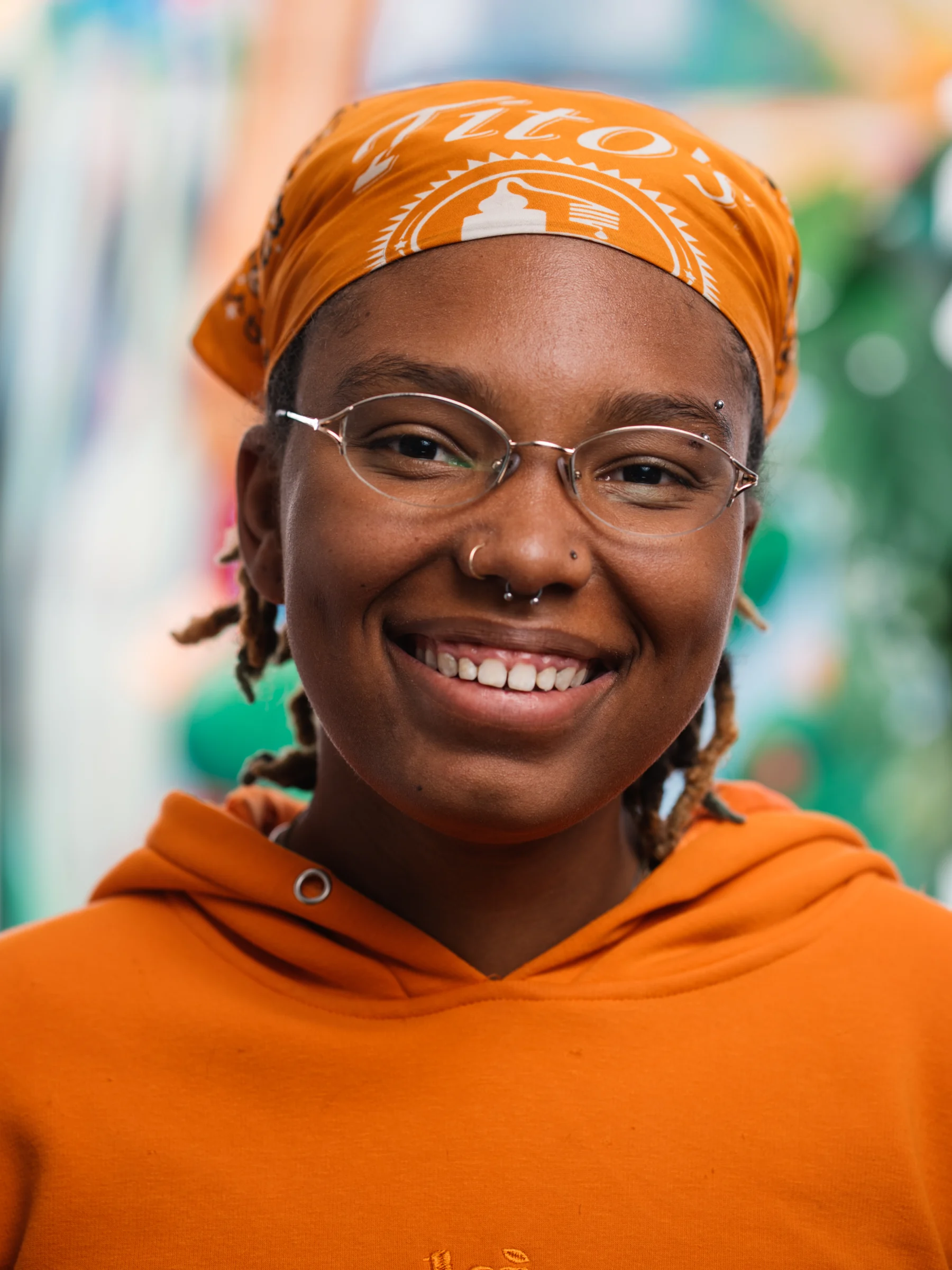 A smiling subject in an orange hoodie and matching head scarf, wearing glasses.