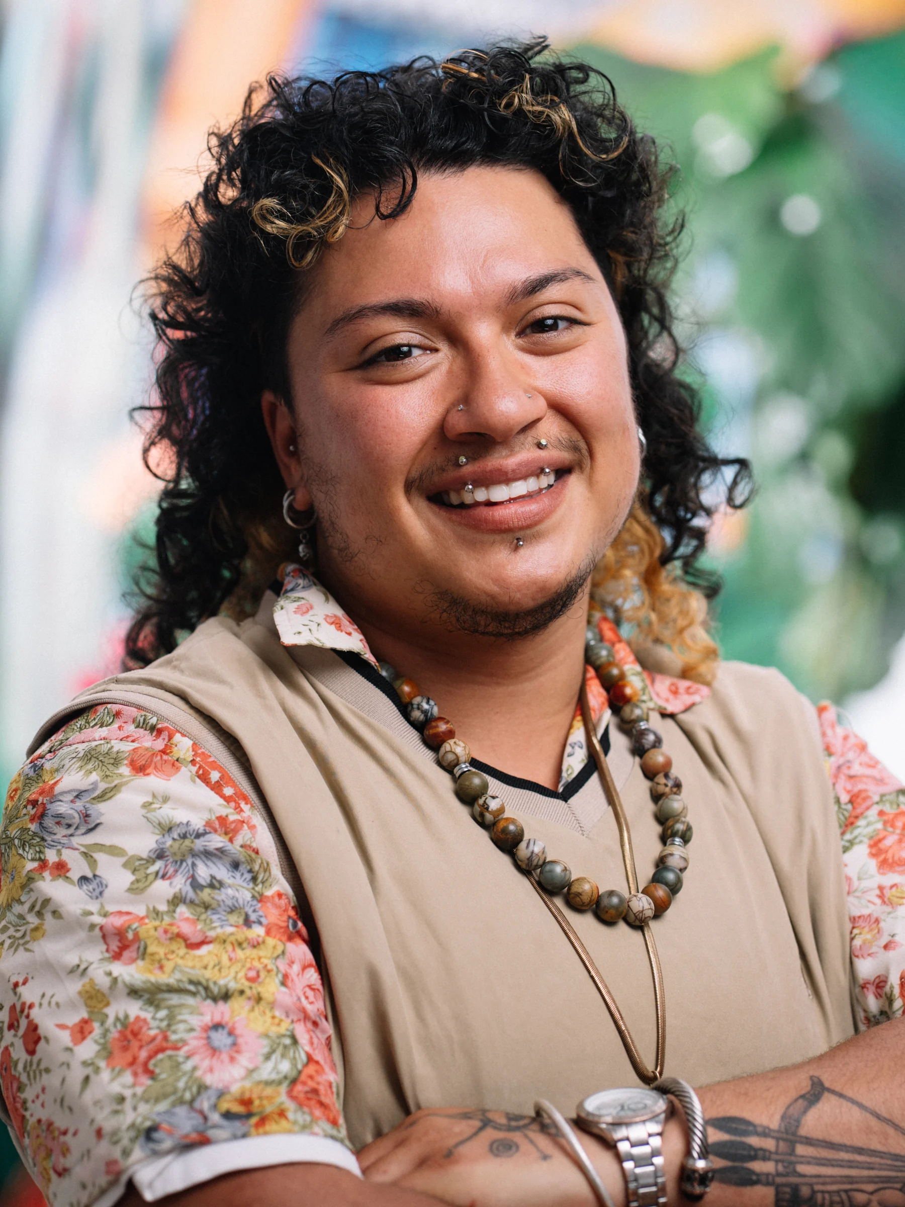 A smiling subject in a floral shirt and tan vest poses with arms folded.
