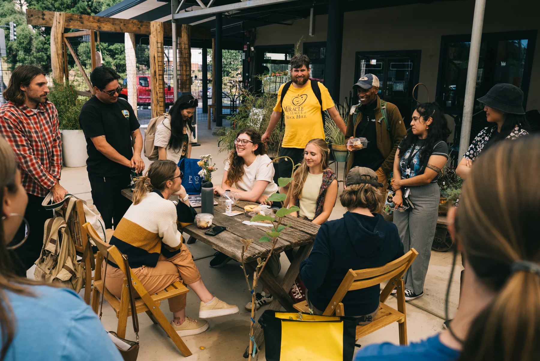 A group gathers around a wooden table, talking and smiling beneath the nursery canopy.