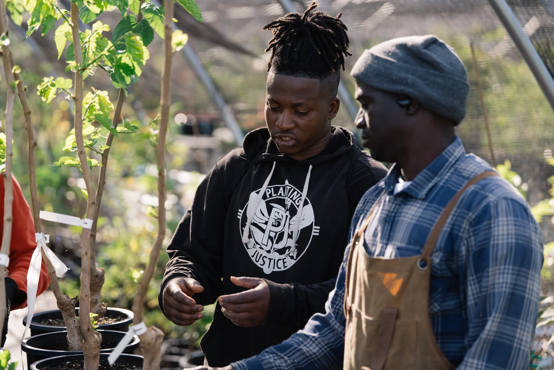 Two people talk beside rows of potted trees at Planting Justice, one gesturing while the other listens.