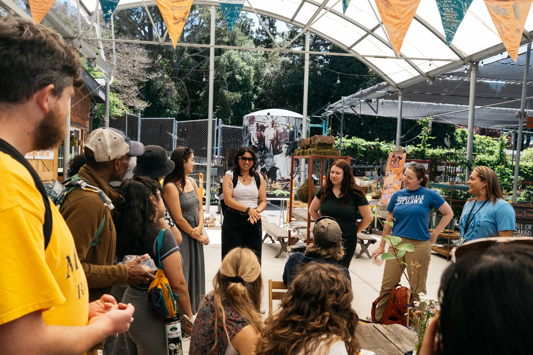 A group listens and talks beneath a greenhouse frame decorated with bunting at the nursery.