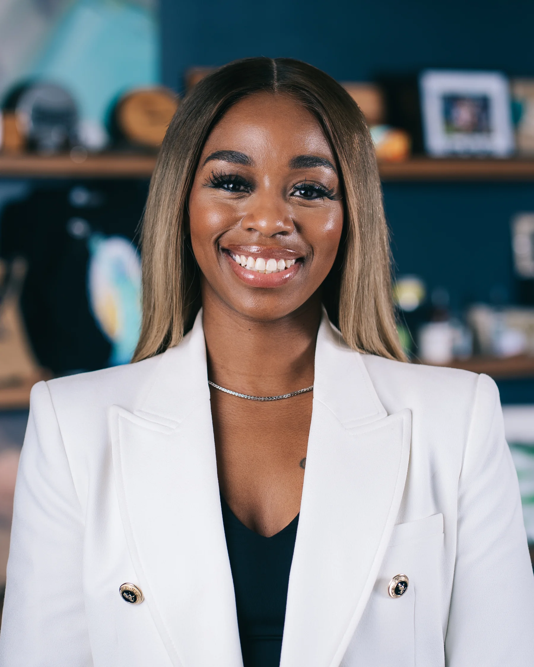 A smiling participant in a light blazer photographed in front of dark teal shelving.