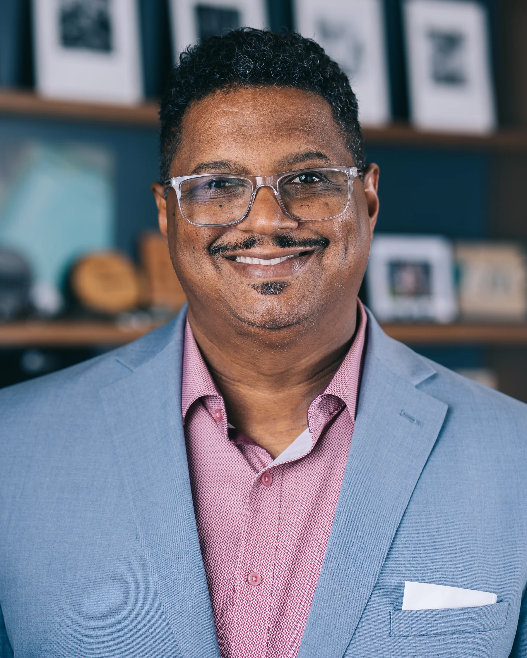A smiling participant in a light blue suit and pink shirt, wearing glasses.