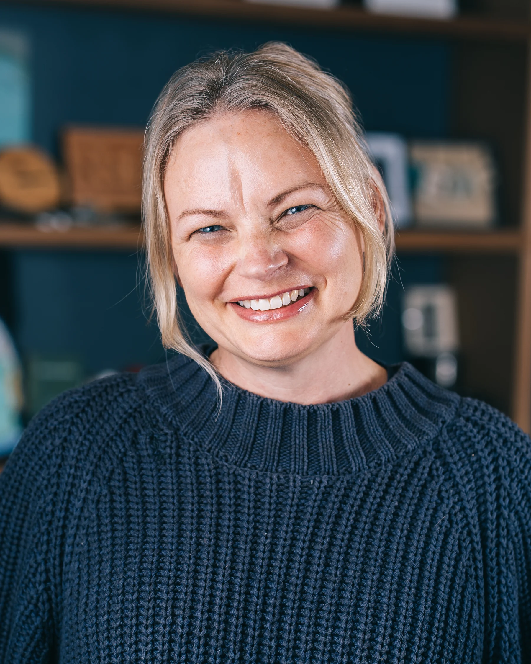 A smiling participant in a dark knit sweater photographed in front of teal shelves.