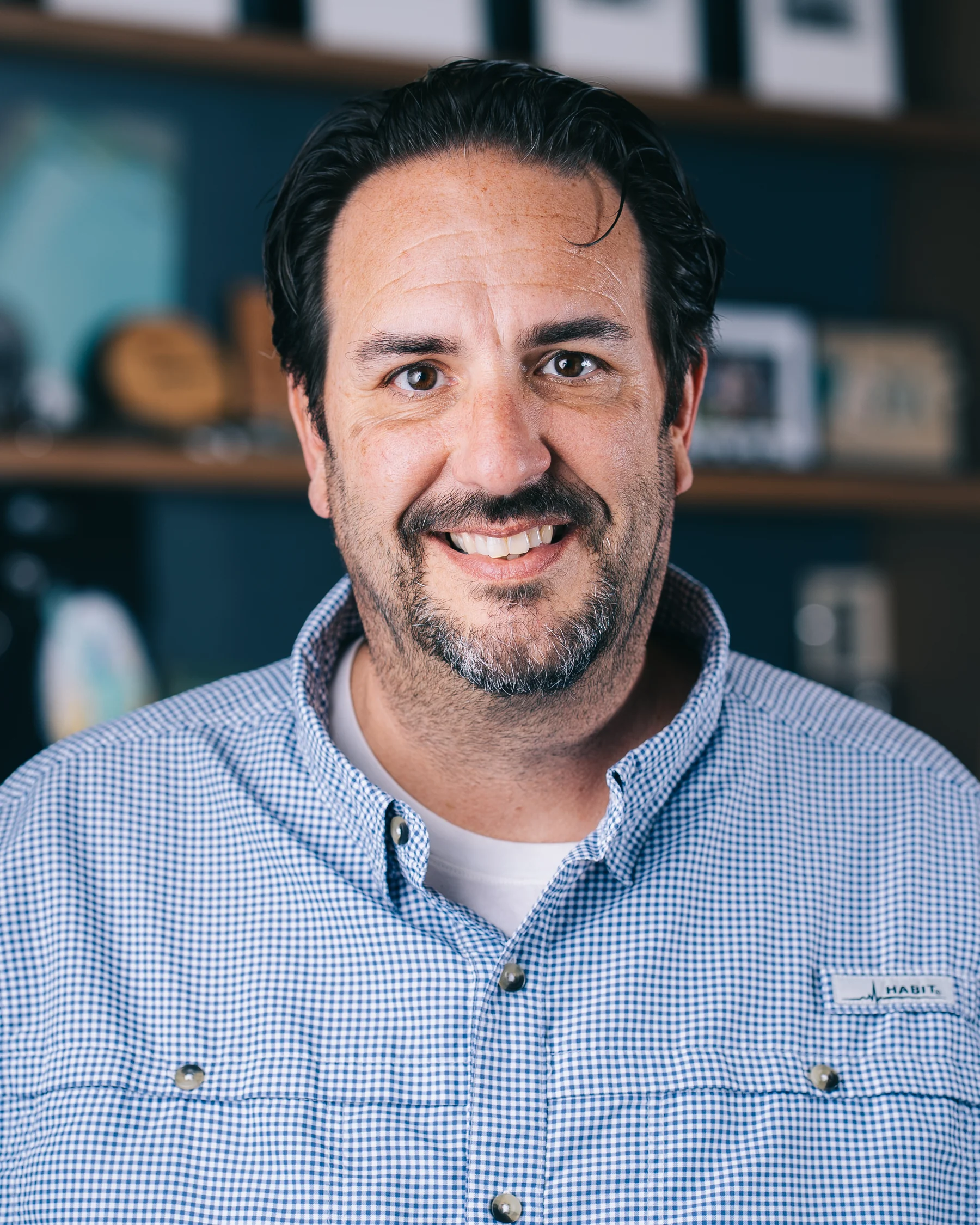 A smiling participant in a blue button-down shirt photographed against teal shelving.