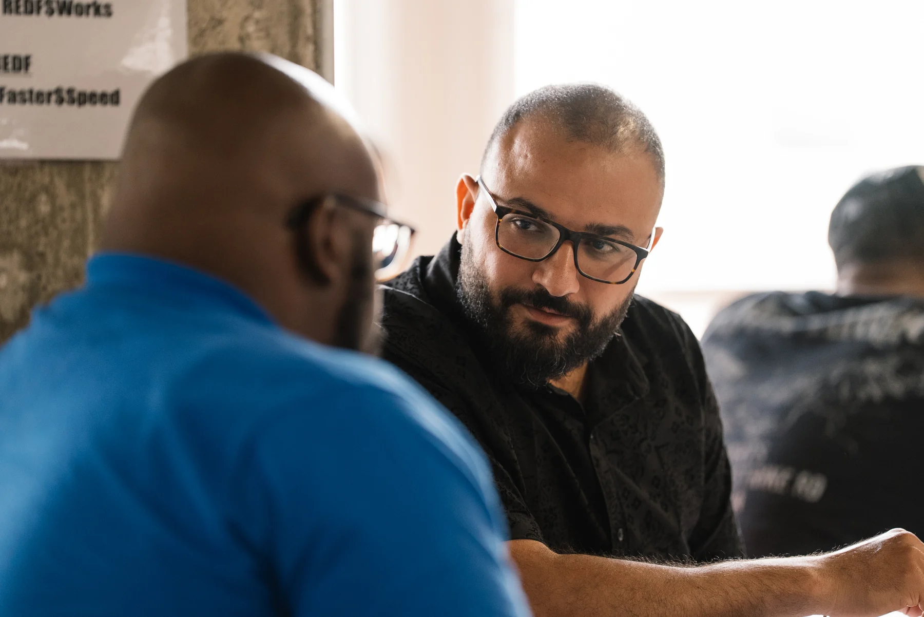 Two Accelerator participants in conversation, with a guest in glasses listening across the table.