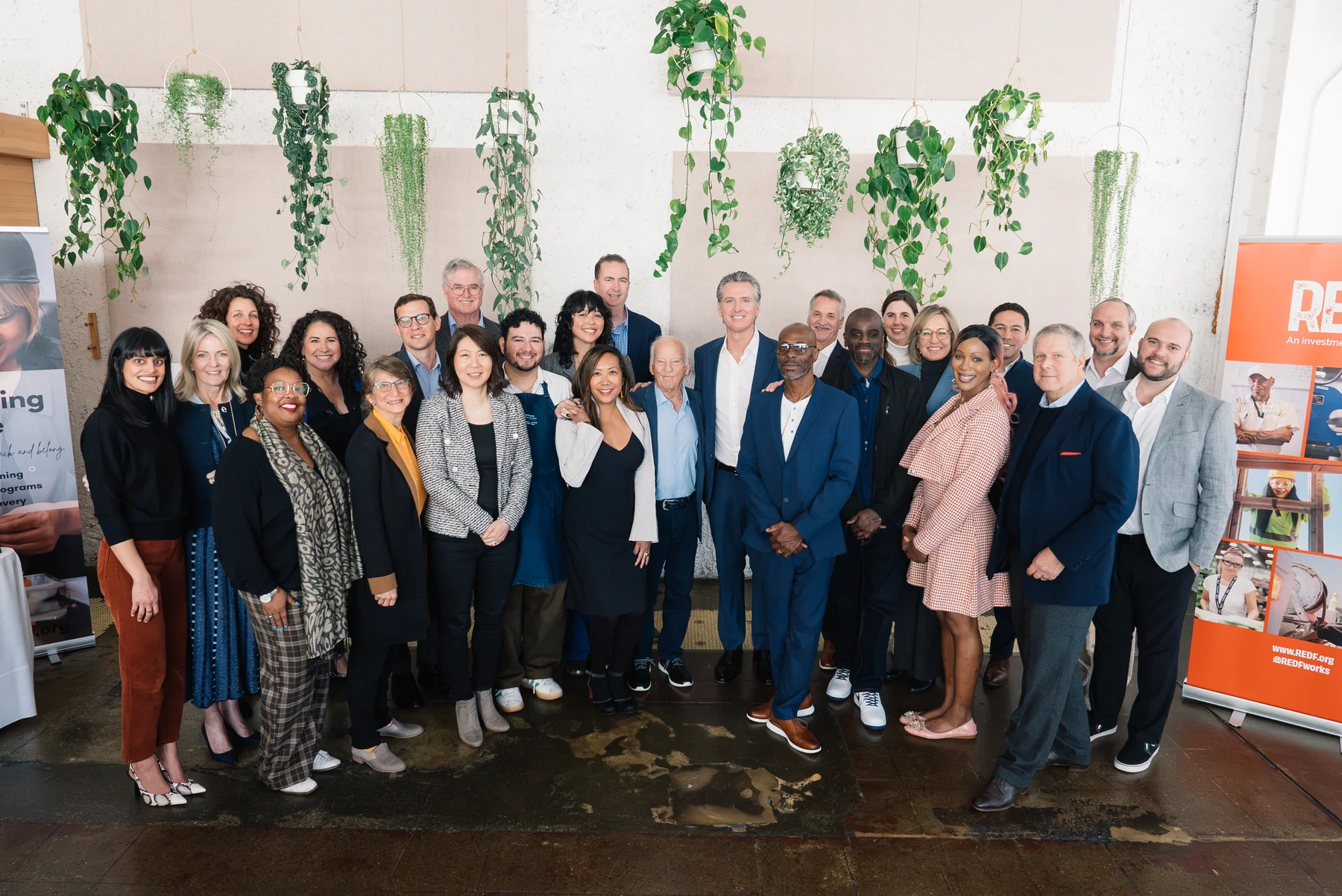 A large group stands together for a portrait in front of a wall of trailing plants.