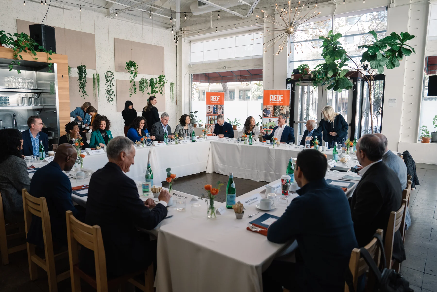 Attendees sit around a U-shaped table in a bright room with large windows and plants.