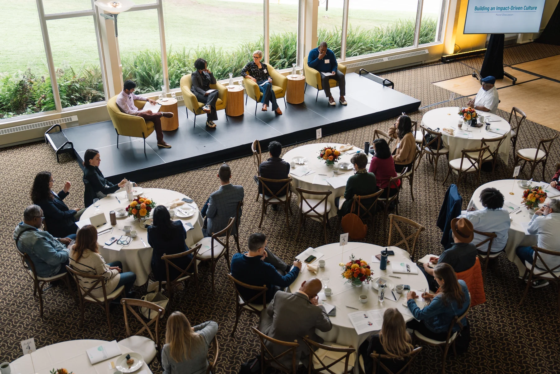 An elevated view of attendees gathered around tables for a panel discussion beside tall garden-facing windows.